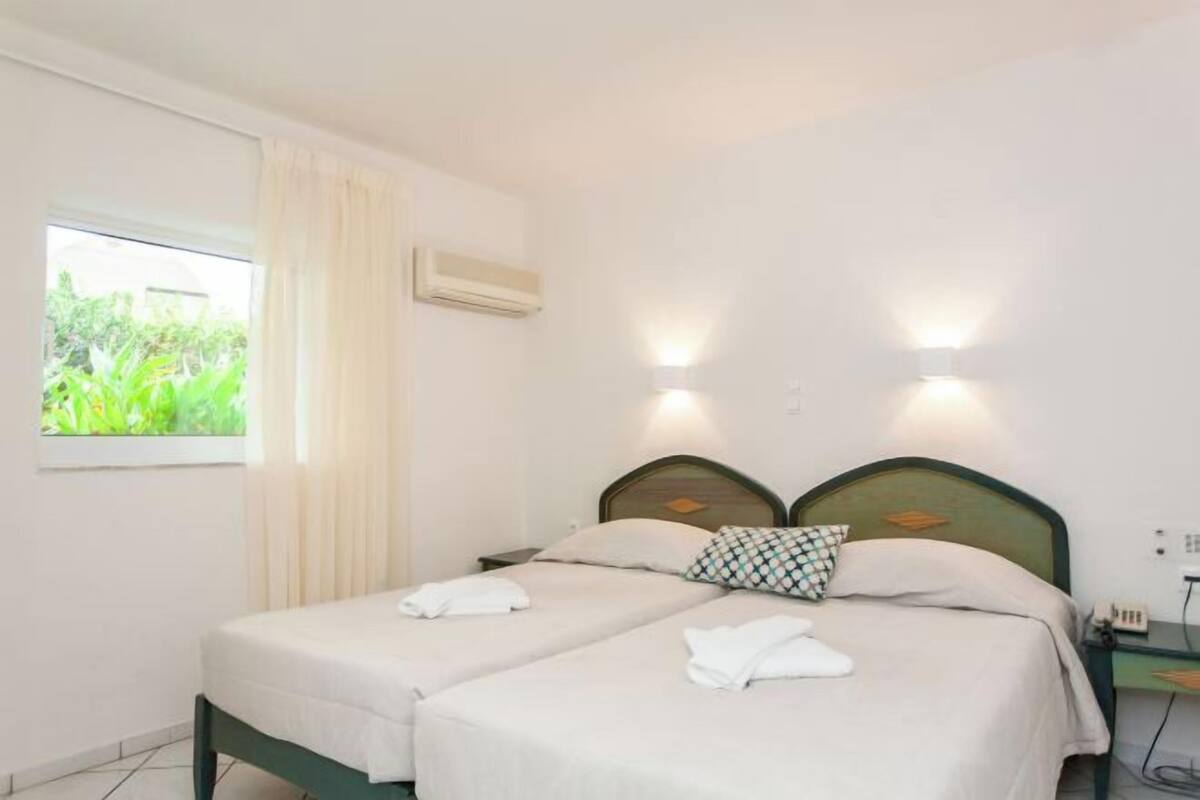 A hotel room with two beds, a window with curtains, an air conditioning unit, and wall-mounted lights.