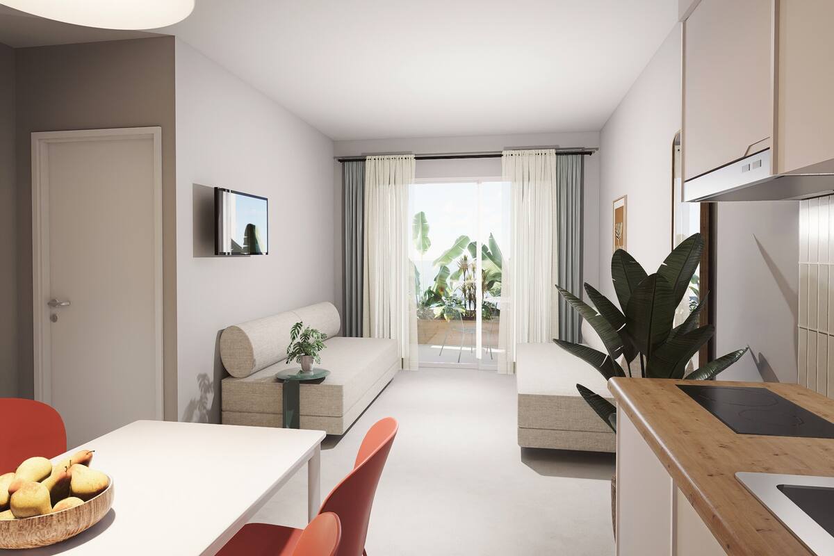 A modern living room with a sofa, dining table, and chairs. There is a kitchen area with a sink and cabinets. A large plant is visible in the corner.