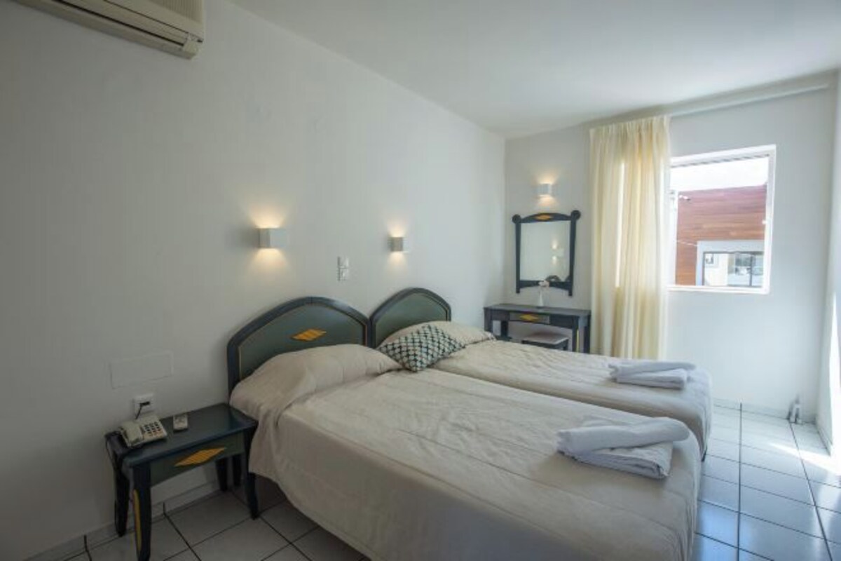 A hotel room with two beds, a bedside table with a phone, a mirror, and a window with curtains.
