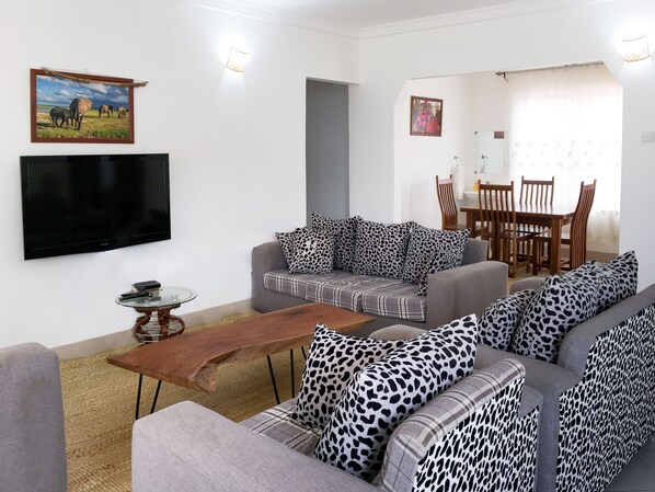 A living room with a sofa set, a coffee table, a flat-screen TV, and a dining area with a table and chairs. - Mama Wilsen House (Arusha)