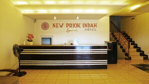 A reception desk at New Priok Indah Hotel with a sign above it, a fan, and a staircase in the background. - New Priok Indah Hotel (Jakarta)