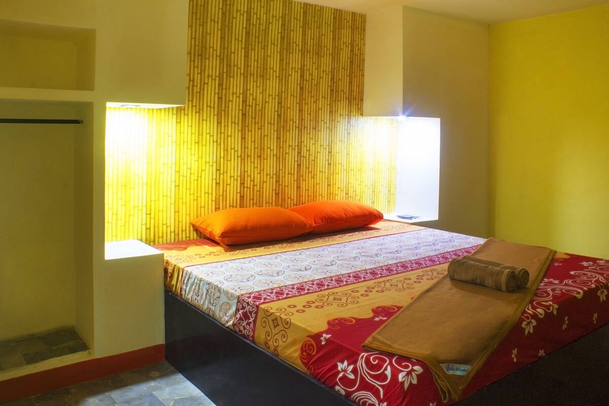 Un lit simple avec une couvre-lit à motifs, un oreiller orange et une couverture enroulée sur une table placée près de la fenêtre.
