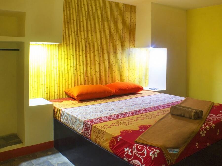 Una cama individual con una colcha estampada, una almohada naranja y una manta enrollada sobre una mesita junto a la ventana.