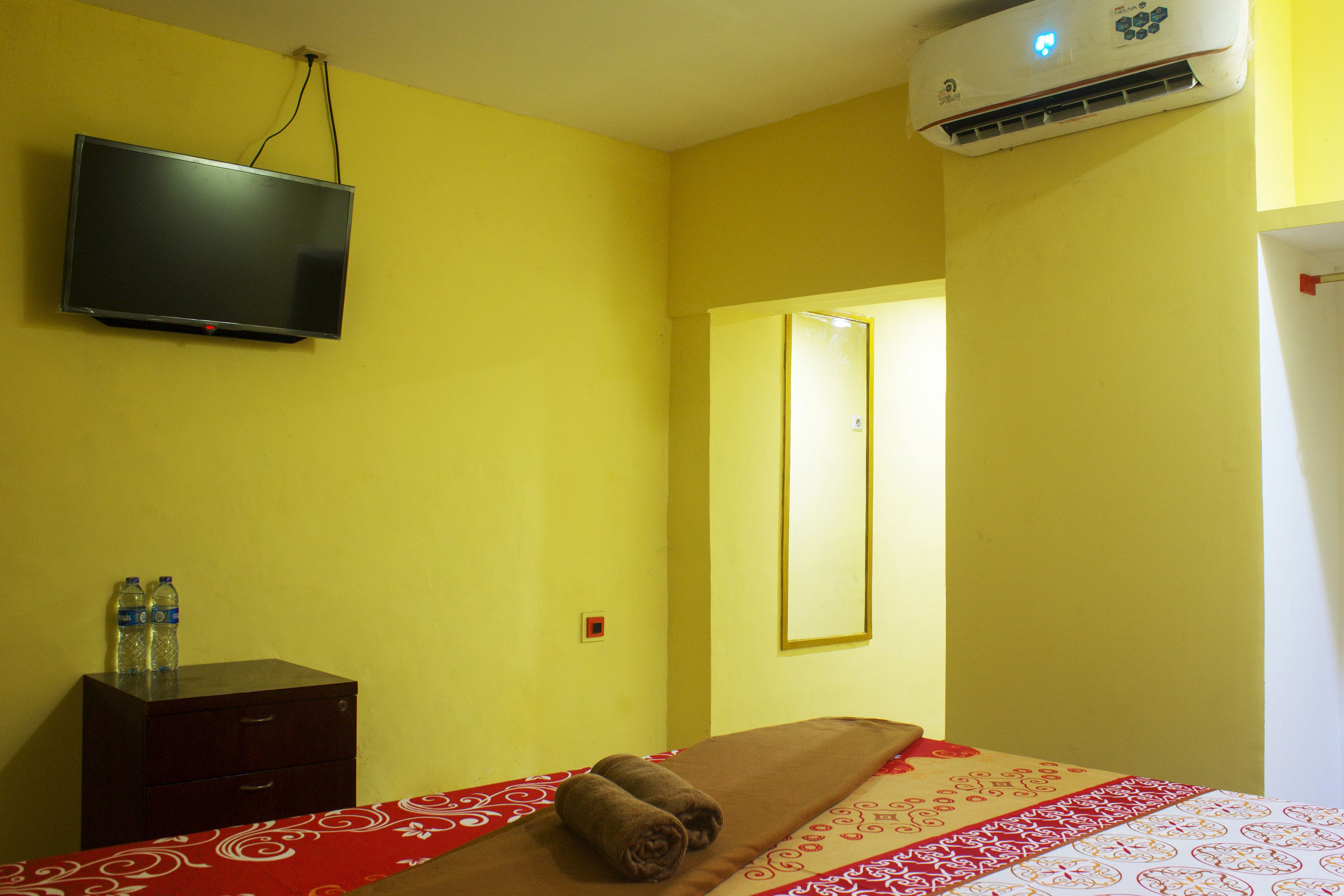 Une chambre d’hôtel avec un lit, un téléviseur fixé au mur, une table de chevet avec deux bouteilles et un appareil de climatisation.