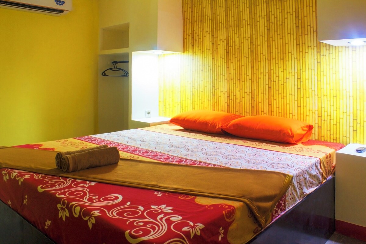 A bed with a red and white patterned bedspread, an orange pillow, and a rolled towel on the side.