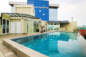 A swimming pool with a glass enclosure, a building with a sign reading "CIRSM KIRANA," and a red-roofed structure in the background. - The Baile (Jakarta)