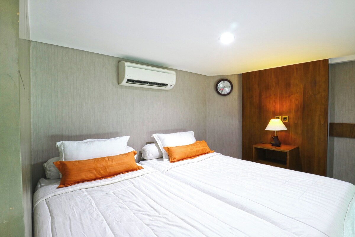 A hotel room with two beds, an air conditioning unit, a clock, and a nightstand with a lamp.