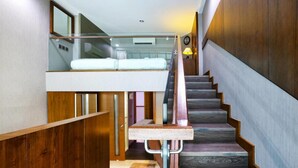 Un escalier moderne avec des marches en bois et une rampe en verre, menant à une chambre mansardée équipée d’un lit superposé.