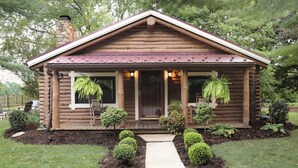 A log cabin with a front porch, a stone chimney, and a small garden with trimmed bushes and a pathway leading to the entrance.