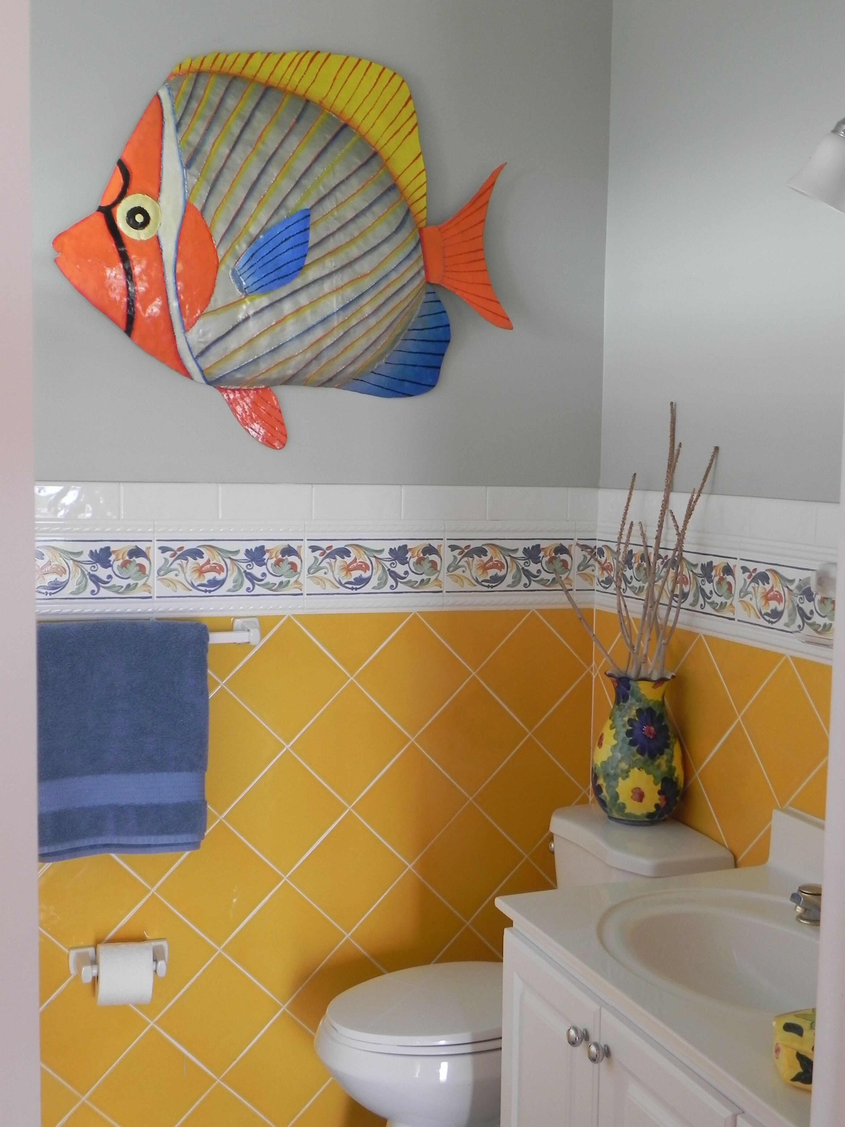 A bathroom with yellow tiles, a white toilet, a blue towel, and a decorative fish on the wall.