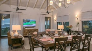 A spacious dining area with a wooden table set for four, flanked by two chairs. A television is mounted on the wall above the table, and there are two floor lamps providing ambient lighting. - Marathon:  Full AC! Pool! Amazing Views! (St. John)