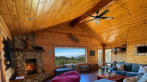 L’intérieur d’un chalet en rondins, avec une cheminée en pierre, un plafond en bois et une grande fenêtre donnant sur le paysage extérieur.