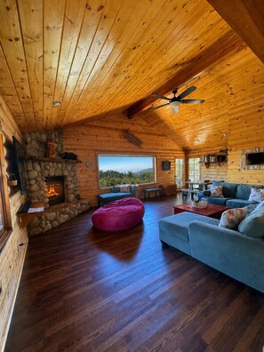 Interior de una cabaña de troncos con chimenea de piedra, techo de madera y un ventanal con vistas al exterior.