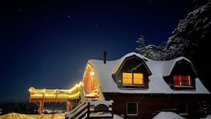 Une cabane recouverte de neige, avec une façade en bois, une terrasse dotée de rampes et des fenêtres illuminées.