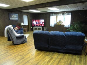 A living room with a blue sofa, a grey armchair, and a television displaying a sports event. - 4BR Home w/ Country Setting & Farmland Views (Lancaster, PA, USA) (Bird in Hand)