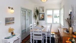 A dining area with a wooden table, white chairs, a vase with flowers, and a view of the outdoors through large windows.