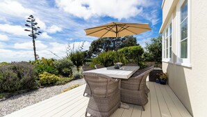 An outdoor patio with wicker furniture and a table under a large umbrella, surrounded by lush greenery and a clear sky. - Your personal ocean-front oasis (Haumoana)