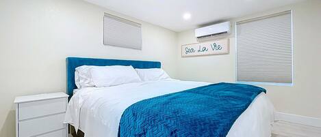 Un dormitorio con cama, mesita de noche, aire acondicionado instalado en la pared y ventana con persianas.