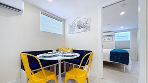 Un pequeño comedor con una mesa redonda, dos sillas amarillas y un cojín azul. En la habitación contigua, hay una cama con cubrecama azul.