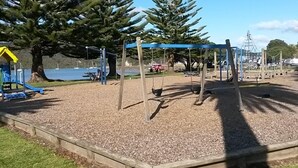Un parque infantil con columpios y un tobogán, rodeado de árboles y una masa de agua.