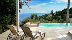 A poolside area with two lounge chairs and a small table, overlooking a clear blue sea and a mountainous landscape.