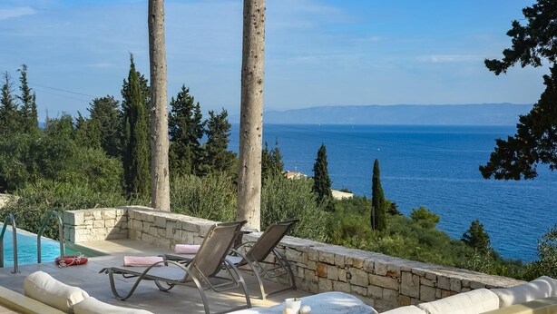 A poolside area with lounge chairs and a stone wall, overlooking a body of water and distant mountains.