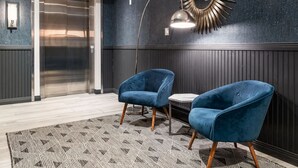 Un vestíbulo moderno con un ascensor grande, dos sillones azules, un tapete estampado y un espejo con diseño de sol en la pared.