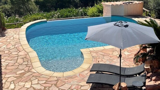 Une piscine bordée de pierres, entourée de chaises longues et d’un parasol, offrant une vue panoramique sur un paysage pittoresque composé de collines et d’une étendue d’eau.