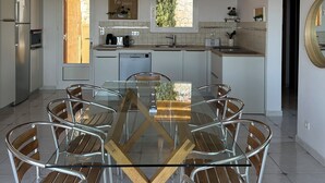 Une cuisine moderne avec des meubles blancs, un plafond en bois et un coin repas comprenant une table en verre et des chaises en métal.