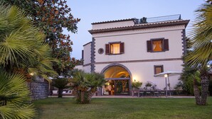A two-story building with a large arched entrance, outdoor seating area, and surrounding greenery. - AMORE RENTALS - Villa Il Pino with Private Pool, Garden, BBQ, Parking (Sant´Agata sui Due Golfi)