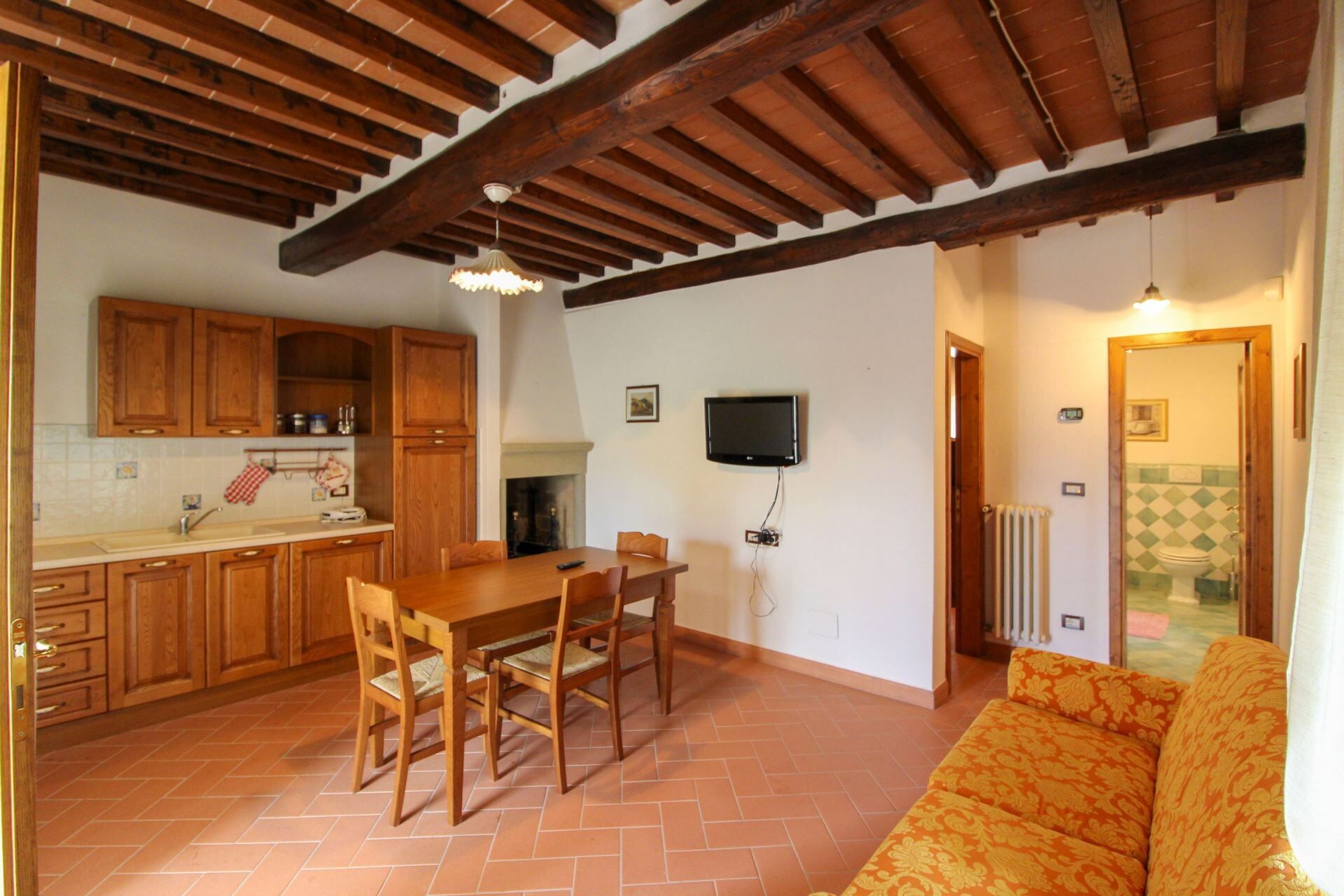 A kitchen with wooden cabinets, a dining table with chairs, a television mounted on the wall, and a patterned sofa in the living area.
