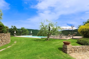 A pool area with a stone wall, a grassy lawn, and outdoor seating with umbrellas.