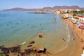 A beach with clear water, rocky shore, and buildings in the background.
