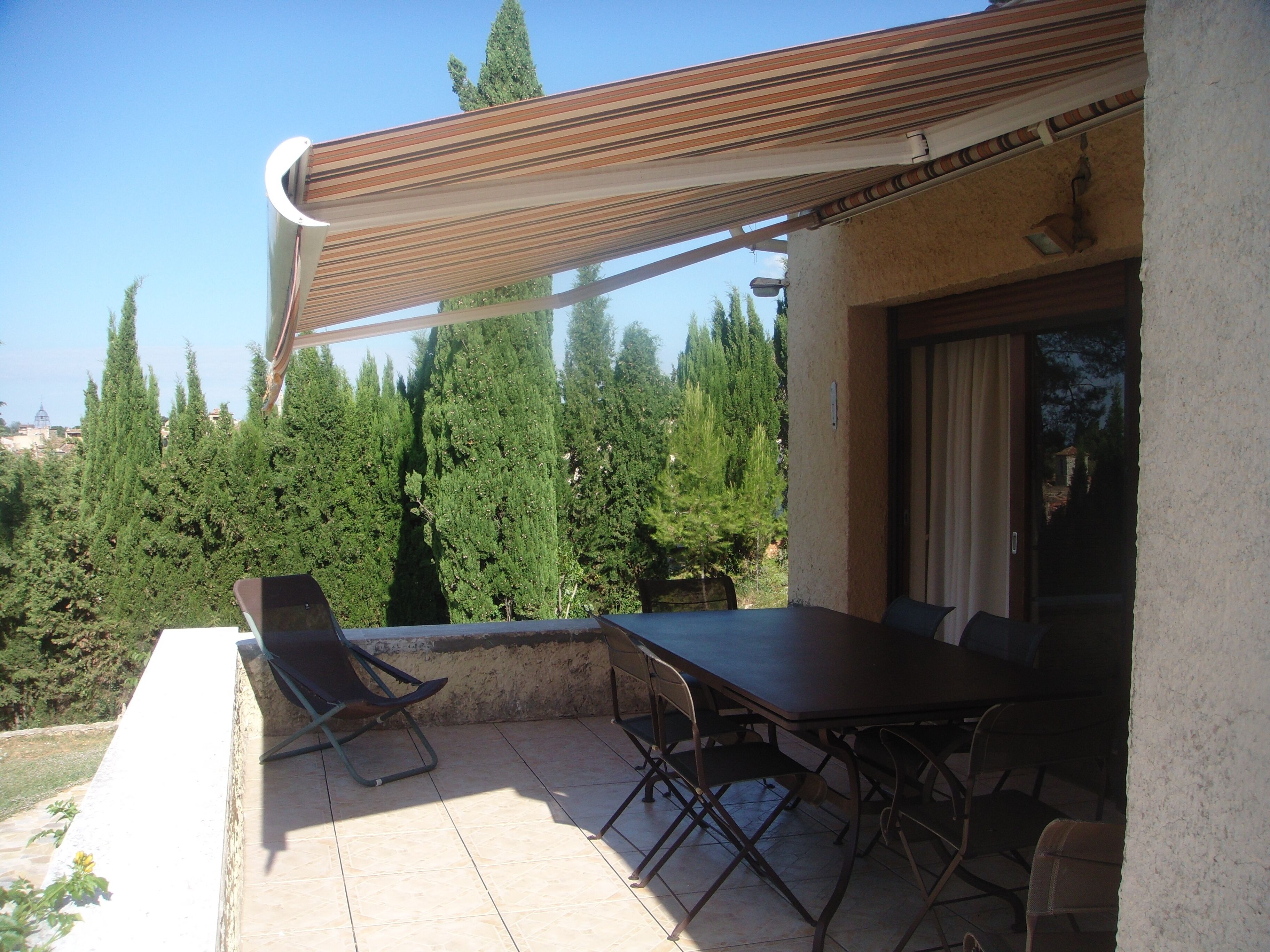 Eine Terrasse im Freien mit Tisch und Stühlen, einem Streifenmarkisenvorzelt und Blick auf hohe Bäume.