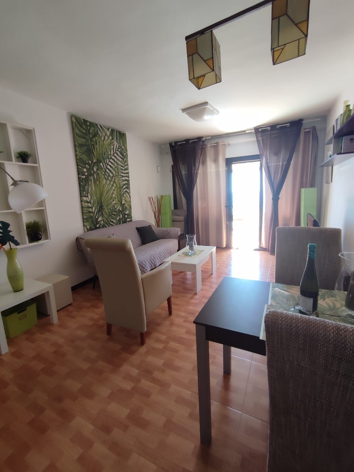 A living room with a sofa, armchair, coffee table, and dining table. There is a bookshelf with decorative items and a large window with curtains.