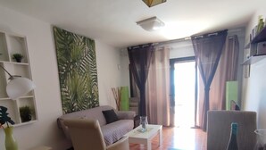 A living room with a sofa, armchair, coffee table, and dining table. There is a bookshelf with decorative items and a large window with curtains. - Quiet apartment near the beach VV-35-3-0002306 (Tías)