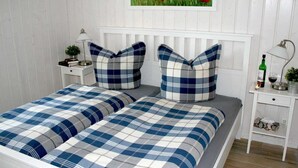 A double bed with blue and white checkered bedding, two matching pillows, a bedside table with a lamp, a small plant, and a framed painting of red flowers on the wall.