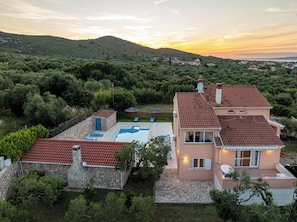 A two-story house with a swimming pool, stone walls, and a view of a hilly landscape.