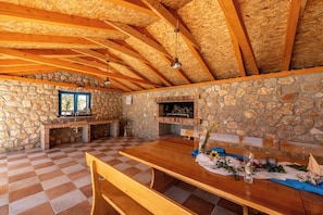 A stone-walled room with a wooden table and benches, a stone fireplace, and a window with blue shutters.