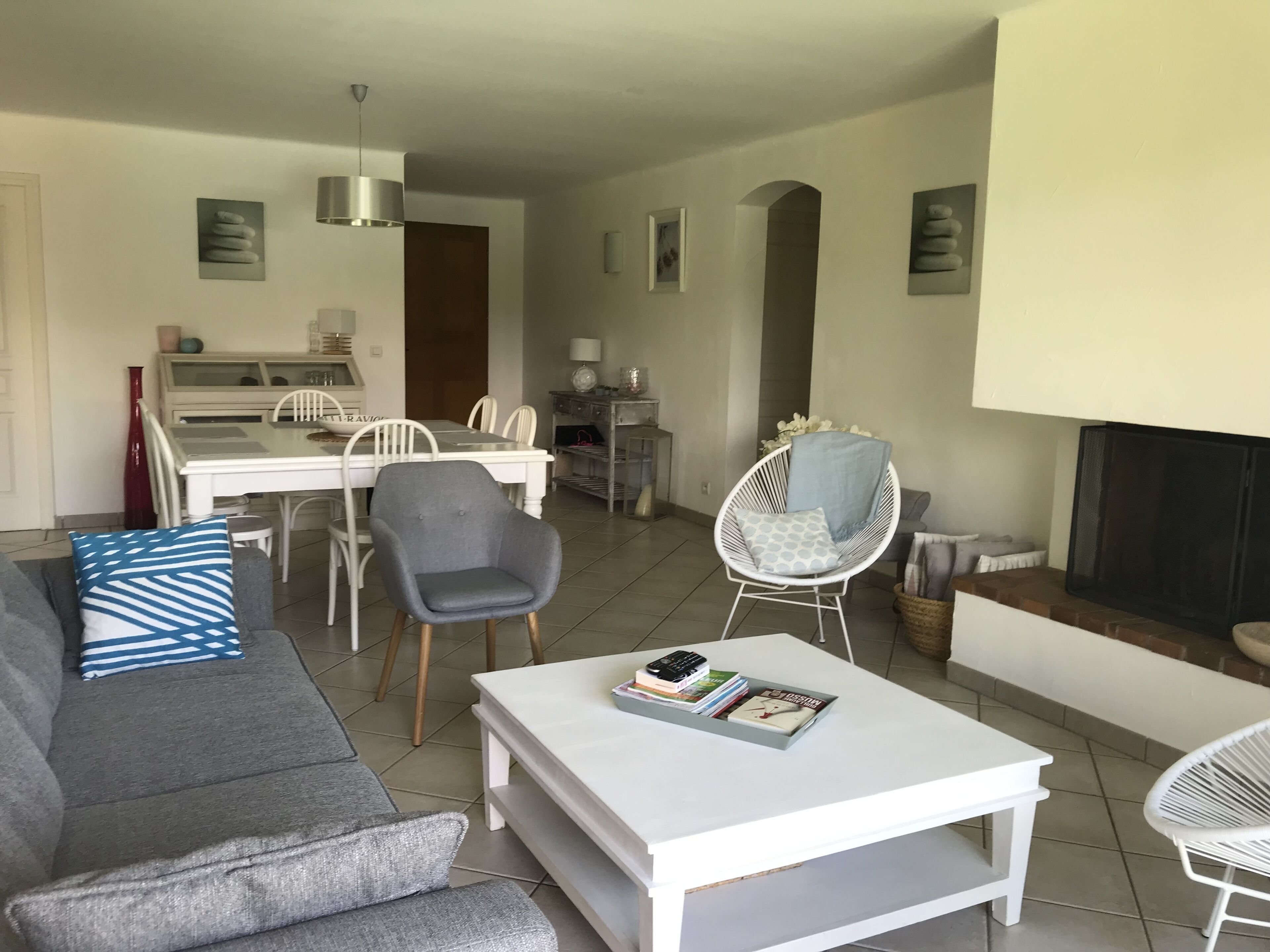 A modern living room with a grey sofa, a white coffee table, and a white chair. There is a dining area with a white table and chairs, and a fireplace with a screen.