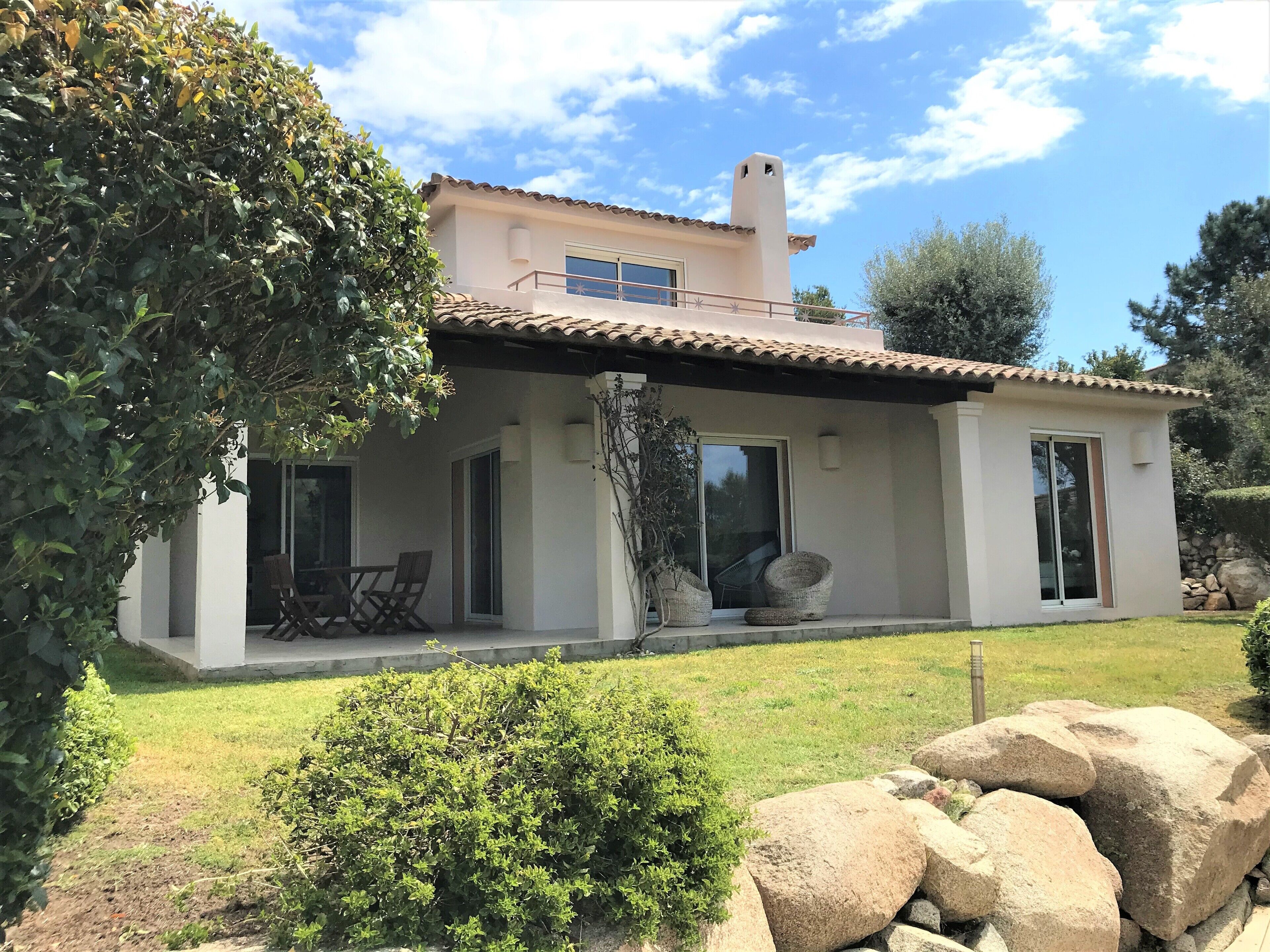 Une maison à deux étages, avec un toit en tuiles, de grandes fenêtres et un patio couvert aménagé avec du mobilier d’extérieur.