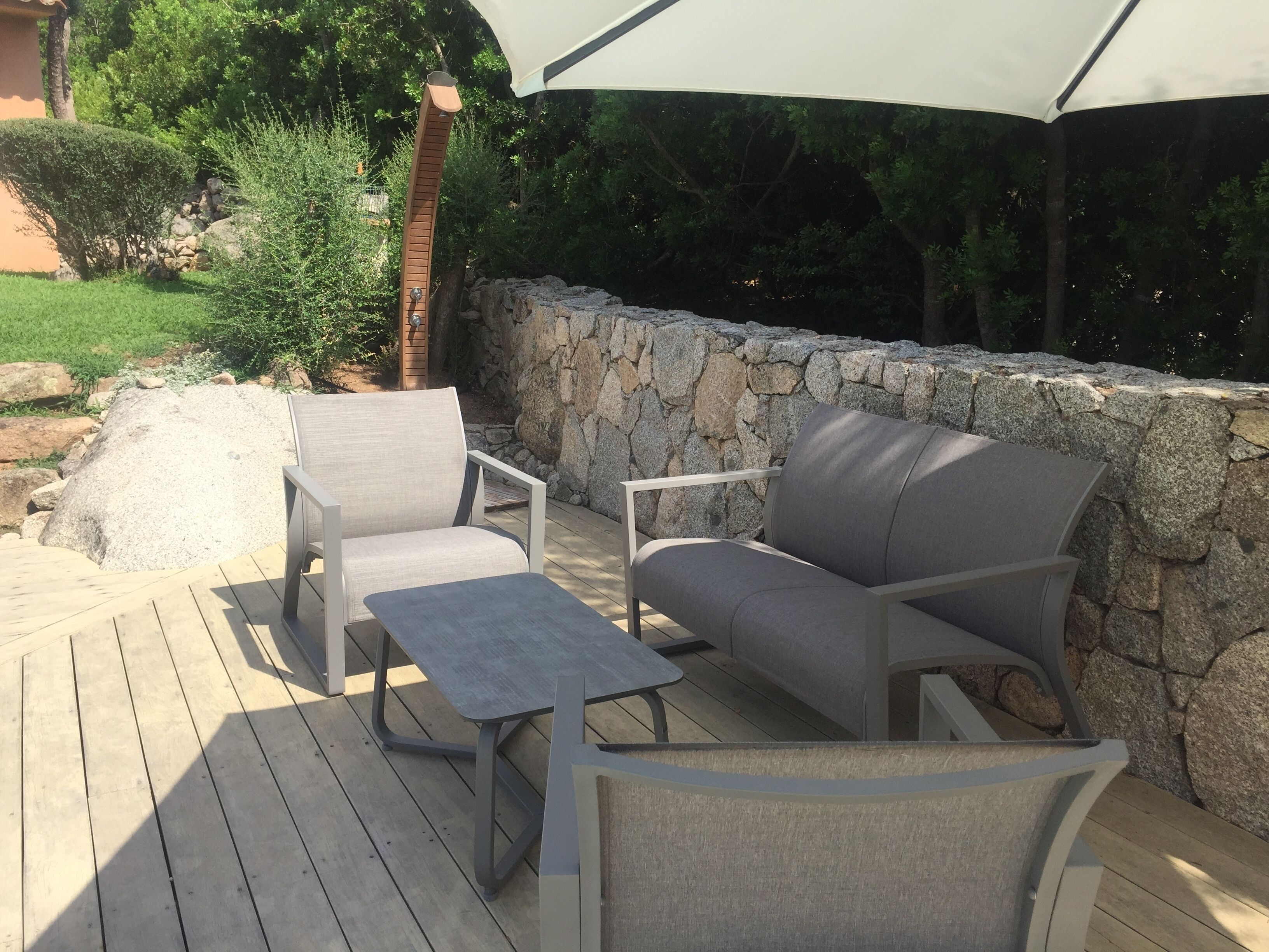 An outdoor patio area with two grey cushioned chairs and a small table, surrounded by a stone wall and a wooden deck.