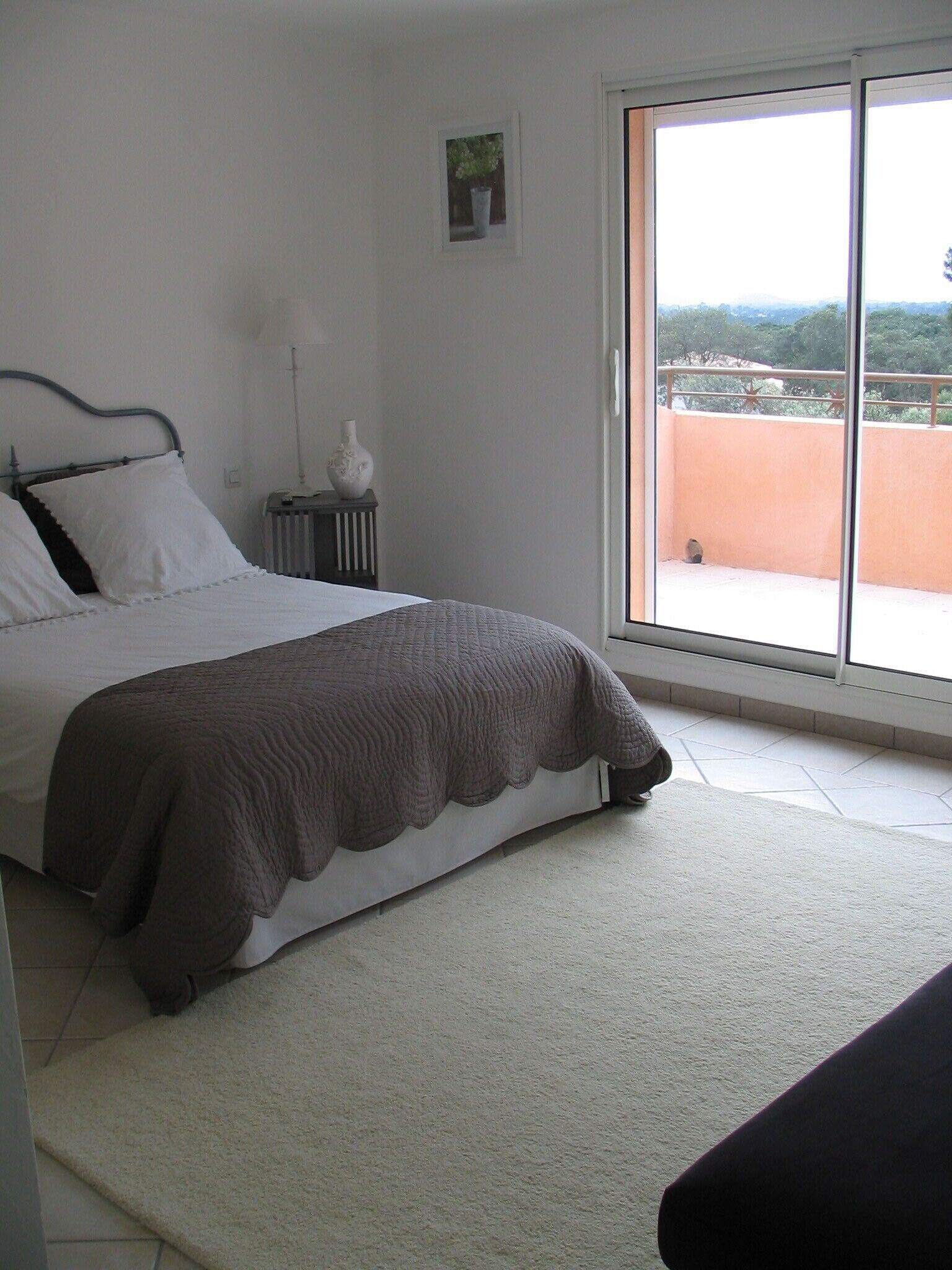 Un dormitorio con cama, un radiador, una mesita con un florero y una ventana que da a vistas del exterior.