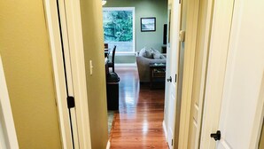 Un couloir doté d’un plancher en bois, de portes blanches et offrant une vue sur un espace de vie comprenant un canapé et une fenêtre.