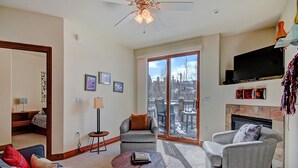 A living room with a fireplace, a television, and a large window with a view of a deck and trees. - Free SkyCard Act-Fitness Center, Gas Fireplace - Highland Greens Lodge 305 (Breckenridge)