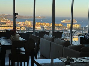 A modern living room with a large window overlooking a cityscape and the sea, featuring a sofa, dining table, and a telescope.