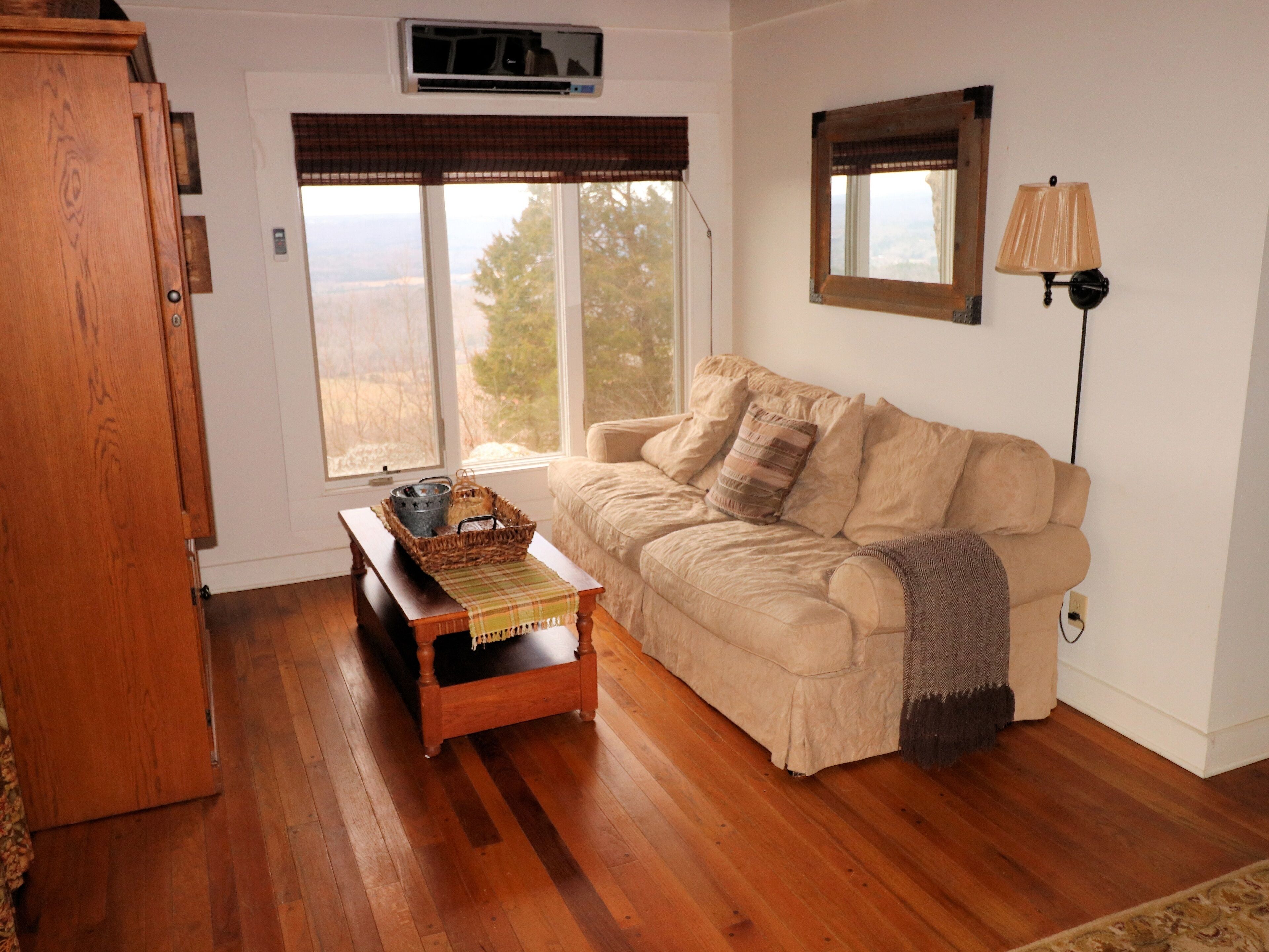 A cozy living room with a wooden floor, a beige sofa, a small wooden table with a basket, and a window with a view of the outdoors.