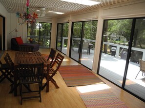 A spacious room with a wooden dining table, chairs, and a colorful rug. Large windows provide a view of the outdoors.