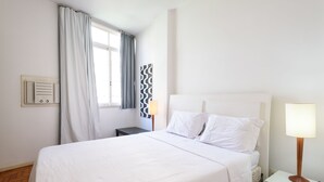 Une chambre à coucher bien rangée, avec un grand lit, une table de chevet avec une lampe, une commode, une fenêtre avec des rideaux et un radiateur fixé au mur.