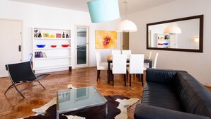 Un salon moderne avec un canapé en cuir noir, une table basse en verre et un coin repas comprenant des chaises blanches et une table en bois. On y trouve une bibliothèque remplie d’objets colorés et un grand miroir accroché au mur.